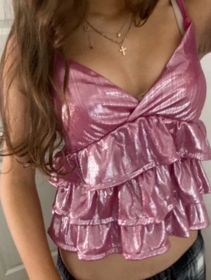 Glittrig rosa volang linnetopp - Säljer en glittrig rosa linnetopp med flera lager volanger och smala axelband. Toppen har omlott-v-ringning framtill och justerbara band. Materialet är glansigt och syntetiskt, perfekt för att sticka ut. Passformen är croppad och toppen är ärmlös.