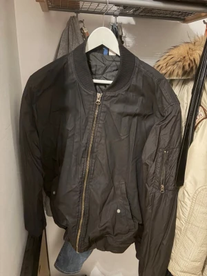 Svart bomberjacka med dragkedja - Säljer en svart bomberjacka med klassisk ribbad krage och muddar. Jackan har en rejäl dragkedja framtill och fickor med tryckknappar samt en extra ficka med dragkedja på ärmen. Perfekt för dig som vill ha en cool och enkel stil.