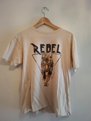 Beige t-shirt med tigertryck från Costes - Cool beige t-shirt från Costes med tryck av en tiger och texten 'REBEL' på bröstet. Klassisk passform med korta ärmar och rund hals. Perfekt för dig som gillar statement-plagg och streetstyle.