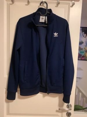 Marinblå Adidas ziptröja XS - Säljer en marinblå ziptröja från Adidas i storlek XS. Tröjan har ståkrage, hel dragkedja och två sidofickor. Adidas-logga i vitt på bröstet. Perfekt för chill eller träning, tillverkad i mjukt syntetmaterial. 
