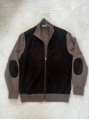 Brun mocka-kofta från Gran Sasso - Snygg brun kofta från Gran Sasso med mocka-paneler framtill och på axlarna. Tillverkad i merinoull och kashmir, med ribbade muddar och svarta mocka-lappar på armbågarna. Dragkedja framtill med läderdetalj. Perfekt för dig som gillar exklusiva material och stilren design.