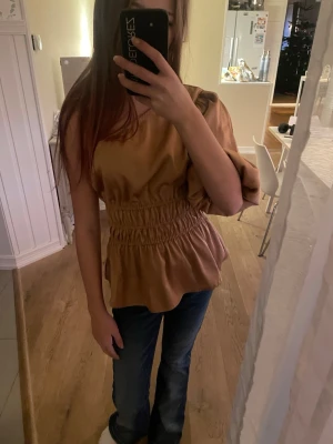 Beige oneshoulder blus med volang - Säljer en beige blus i oneshoulder-modell med en puffig ärm och smockad midja. Toppen har volang nertill och är tillverkad i ett mjukt tyg som ger ett snyggt fall. Perfekt för dig som vill sticka ut lite extra med en unik siluett.