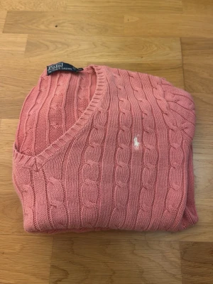 Rosa kabelstickad tröja Polo Ralph Lauren - Kabelstickad tröja från Polo Ralph Lauren i en snygg rosa färg. Klassisk v-ringning och diskret broderad logga på bröstet. Tillverkad i mjuk bomull och har lång ärm. Perfekt för dig som vill ha en stilren och tidlös look.