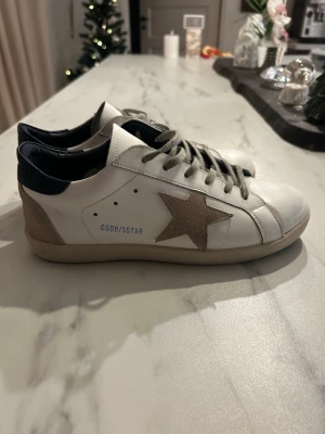 Golden Goose Superstar sneakers - Golden Goose Superstar sneakers i vitt skinn med beige mockadetaljer och en stor stjärna på sidan. Skorna har grå snörning, mörkblå häl och platt sula. Snygg och ikonisk design som sticker ut med sin kontrast mellan färger och material Box ingår. Hör av er vid några frågar. Pris kan diskuteras.