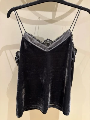 Svart sammets linnetopp med spets - Säljer en svart linnetopp i mjuk sammet med tunna axelband och dekorativ spets längs v-ringad halsringning. Spetsen har ett detaljerat blommönster som ger en lyxig känsla. Toppen är ärmlös och har en loose passform, perfekt för att styla med jeans eller kjol.