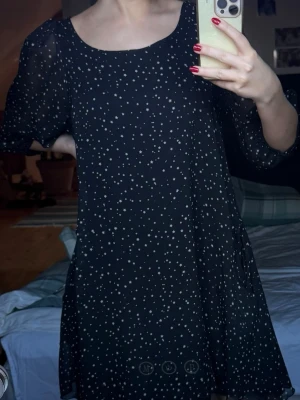 Svart kortklänning med stjärnmönster - Säljer en svart kortklänning med vita stjärnor över hela tyget. Klänningen har puffiga, transparenta kortärmar och en rundad halsringning. Materialet känns lätt och luftigt, perfekt för dig som gillar en lekfull och trendig look.