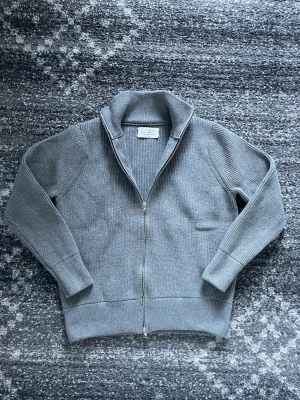 Grå stickad kofta från Maison Margiela - Säljer en grå stickad kofta från Maison Margiela med hel dragkedja och hög krage. Tröjan har ribbade muddar och långärmade ärmar, perfekt för kyliga dagar. Klassisk och stilren design som funkar till många olika outfits.