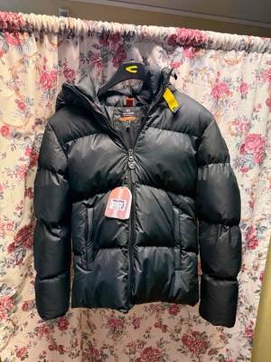 Svart pufferjacka från Parajumpers - Svart pufferjacka från Parajumpers i storlek M. Jackan har en schysst vadderad look, huva och dragkedja framtill. Materialet är en mix av bomull och polyester, vilket gör den både mjuk och slitstark. Perfekt för kalla dagar och har en cool gul detalj vid kragen.