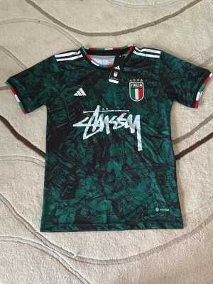 Italien x Stüssy x Adidas t-shirt - Grym grön t-shirt från Adidas i samarbete med Stüssy och Italiens landslag. Har coolt konstinspirerat mönster, vita Adidas-ränder på axlarna, broderad Italiens badge och Stüssy-logga framtill. Ribbad krage och ärmslut i Italiens färger. Slim fit och tillverkad i polyester.