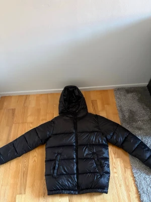 Svart pufferjacka från Grunt XL - Säljer en svart pufferjacka från Grunt i storlek XL. Jackan har huva, två snedställda fickor framtill och dragkedja. Materialet är glansigt och känns riktigt mjukt, perfekt för kalla dagar. Enkel och clean design som passar till det mesta.
