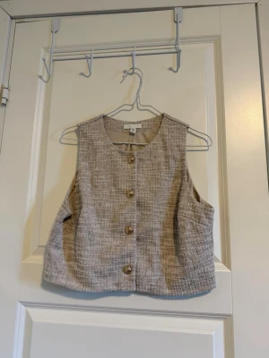 Beige väst med knappar från H&M - Säljer en beige väst från H&M med snyggt rutigt mönster och 4  knappar framtill. Västen har en rak och lite loose passform. Perfekt att styla över en t-shirt eller skjorta för en chill look.