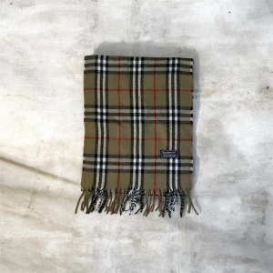 Burberry halsduk - Säljer en klassisk rutig halsduk från Burberry i beige, svart, vit och röd. Halsduken är tillverkad i 100% lammull. Halsduken är perfekt nu under vintern för att kunna hålla sig varm och stilren. Tveka inte att höra av dig vid frågor eller funderingar.