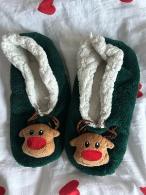 Juliga tofflor med renmotiv - Mysiga tofflor i mörkgrönt och beige med vit fluffig insida. På ovansidan finns ett gulligt renansikte med röd nos och bruna horn. Perfekta för kalla vinterdagar och julmys! Storlek 36.