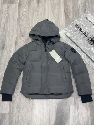 Grå dunjacka från Canada Goose - Säljer en grå dunjacka från Canada Goose med huva och svart logotyp på ärmen. Jackan har quiltad design, dragkedja framtill och två sidofickor. Perfekt för kalla dagar och har en clean, modern look som funkar till streetwear.