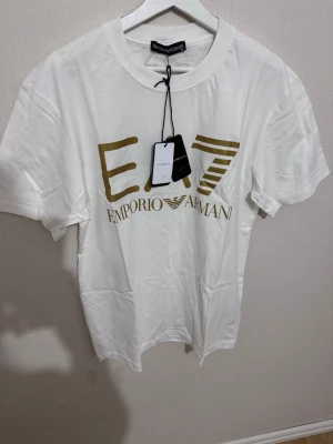 Vit EA7 Emporio Armani t-shirt - Snygg vit t-shirt från EA7 Emporio Armani med guldigt tryck på bröstet. Klassisk rund hals och korta ärmar. Perfekt för dig som gillar stilrena märkesplagg med en exklusiv touch. Materialet är mjuk bomull som känns skönt mot huden.Fraktar samma dag eller nästa✅hör av dig via frågor!