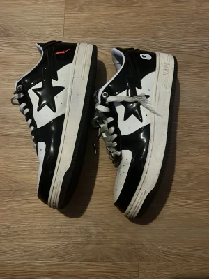 Svarta och vita Bape Sta sneakers - Säljer ett par Bape Sta sneakers i svart och vitt med den ikoniska stjärnan på sidan. Skorna har snörning, perforerad tå och platt sula. Märket syns tydligt på plösen och hälen. Materialet är blankt skinn och de har en rund tå. Perfekt för dig som gillar streetwear och vill sticka ut. Osäker om dessa är äkta !!!