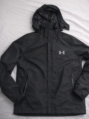 Svart vindjacka från Under Armour - .Varm och bekväm Under Armour-jacka i slitstarkt, vattenavvisande material. Stilren design med huva och dragkedjefickor – perfekt för kyliga dagar. Mycket bra skick.