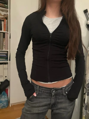 Svart zip-kofta med lång ärm - Slimmad svart kofta med dragkedja framtill och långa ärmar. Modellen är kroppsnära och har en enkel, stilren look som passar till jeans eller kjol. Materialet känns mjukt och stretchigt, perfekt för lager-på-lager. Snygg att ha öppen eller stängd beroende på stil.