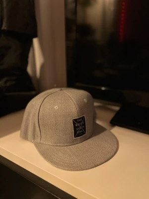 Keps - Bilderna visar en grå keps med rak skärm och justerbar snapback, samt en patch med texten 'The Good Vibes Only' framtill. Ingen väst syns på bilderna.