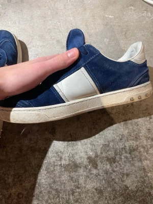 Valentino Garavani blå sneakers - Snygga sneakers från Valentino Garavani i mörkblå mocka och vitt skinn. Skorna har snörning, rund tå och platt sula med nitar på hälen. Yttersulan har ett coolt kamouflagemönster och logotyp. Perfekta för dig som gillar exklusiva streetwear-vibes.
