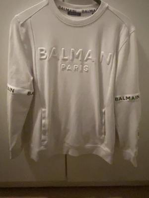 Vit sweatshirt från Balmain Paris - Säljer en vit sweatshirt från Balmain Paris med upphöjd logotyp på bröstet och tryck på ärmarna. Tröjan har rund hals, långa ärmar och ribbade muddar. Perfekt för dig som gillar exklusiva märken och stilren design.
