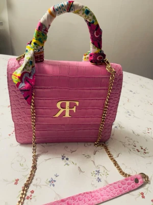 Rosa handväska med guldkedja Roberto Fabiani - Säljer en snygg rosa handväska från Roberto Fabiani med krokodilpräglat mönster och guldfärgad kedja. Handtaget är inslaget i ett färgglatt tyg med blommigt mönster. Väskan har en stilren rektangulär form och ett stort RF-emblem i guld på framsidan. Perfekt för dig som gillar färg och detaljer!