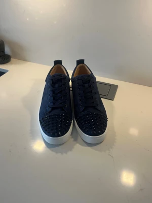 Mörkblå sneakers från Christian Louboutin - Säljer ett par mörkblå sneakers från Christian Louboutin med coola nitar på tån och klassisk röd sula. Skorna har snörning, rund tå och är gjorda i mocka med vita platta sulor. Insidan är beige och har Louboutin-loggan i guld.