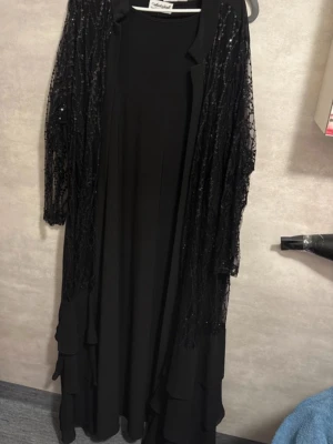 Svart långklänning med paljettdetaljer. Abaya  - Svart långklänning från Andy Gold med långärmade meshpartier täckta av svarta paljetter. Klänningen har volangdetaljer nedtill och ett elegant, flytande fall. Perfekt för dig som gillar glam och vill sticka ut med glittriga detaljer.