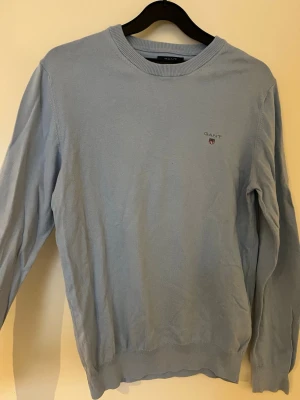 Ljusblå långärmad tröja från GANT - Snygg ljusblå långärmad tröja från GANT med rund hals och diskret logga broderad på bröstet. Tröjan är i mjuk bomull och har ribbade muddar vid hals, ärmslut och nederkant. Perfekt för dig som gillar stilrena och klassiska plagg.