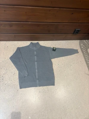 Grå stickad kofta från Stone Island - Säljer en grå stickad kofta från Stone Island med hel dragkedja och ribbad struktur. Klassisk logotyp-patch på vänster ärm och hög krage. Perfekt för dig som gillar stilrena och snygga plagg med street-vibe.