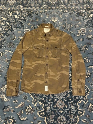Camouflage skjorta Abercrombie & Fitch - Säljer en snygg camouflage-mönstrad skjorta från Abercrombie & Fitch i storlek S. Skjortan har två bröstfickor med knappar, klassisk krage och långa ärmar. Materialet är mjuk bomull och passformen är muscle fit. Perfekt för dig som gillar streetwear och utility-vibe.
