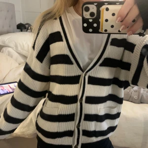 Randig kofta från Lindex i svart och vitt - Säljer en snygg och mysig kofta från Lindex i storlek M. Koftan är stickad med breda svarta och vita ränder, har v-ringad hals och knäpps med knappar framtill. Perfekt att slänga på sig över en t-shirt för en chill look.