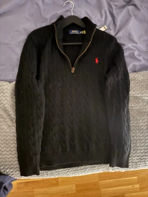 Svart stickad tröja Polo Ralph Lauren - Svart kabelstickad tröja från Polo Ralph Lauren med röd broderad logga på bröstet. Tröjan har half zip med snyggt läderdetalj på dragkedjan och ribbade muddar. Perfekt för dig som gillar klassisk stil med en modern touch. Använt max 2 gånger inga skador alls!