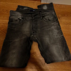 Grå jeans från G-Star RAW, stl 32/32 - Snygga grå jeans från G-Star RAW med slitna detaljer och diskret logga på fickan. Jeansen har klassisk femficksdesign, raka ben och är tillverkade i mjukt denimtyg. Perfekta för dig som gillar en avslappnad men trendig look.