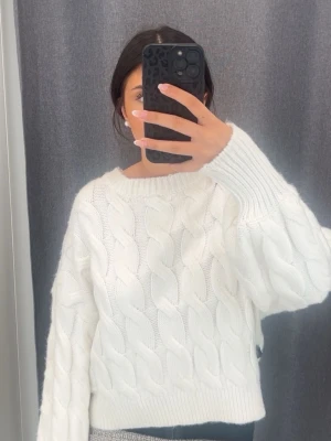 Vit stickad tröja med snörning i ryggen - Säljer en vit, chunky stickad tröja med grovt flätmönster och snygg snörningsdetalj i ryggen. Tröjan har rund halsringning och långa ärmar med breda muddar. 