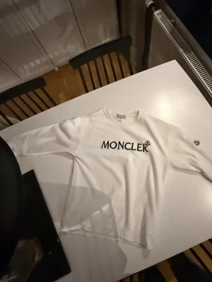 Vit sweatshirt från Moncler, storlek L - Säljer en vit sweatshirt från Moncler i storlek M-L. Tröjan har svart Moncler-tryck på bröstet och en rund hals. Långa ärmar med Moncler-logga på vänster ärm. Tillverkad i mjuk bomull, perfekt för chill dagar. Enkel och stilren design.