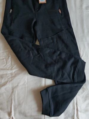 Barn mjukisbyxor strl.140 från Lager 157 - Svarta mjukisbyxor från Lager 157, modell Cooper. Byxorna har dragkedjor vid fickorna och resår i midjan med snörning. Materialet är 63% bomull, 34% polyester och 3% elastan vilket gör dem riktigt sköna och flexibla. Perfekta för chill eller träning.