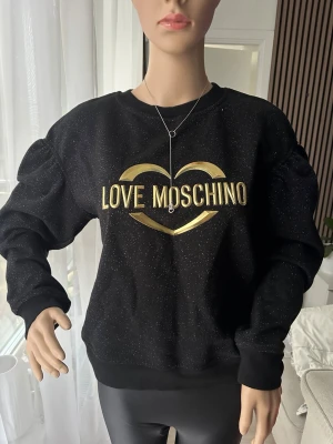 Svart glittrig sweatshirt Love Moschino - Cool svart sweatshirt från Love Moschino med guldigt hjärta och logga framtill. Tröjan har glittriga detaljer i tyget och puffiga ärmar för en trendig look. Rund halsringning och mjukt material som känns skönt mot huden.