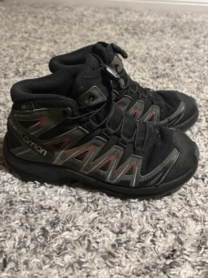 Salomon XA Pro svarta kängor - Salomon XA Pro kängor i svart med grå och röda detaljer. Skorna har snörning med snabbspänne, robust platt sula och meshpartier för extra ventilation. Perfekta för utomhusaktiviteter och äventyr. 