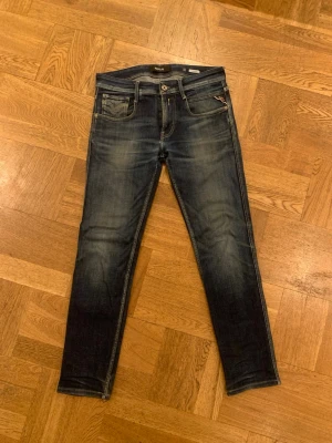 Replay Anbass blå jeans, storlek 30 - Säljer ett par Replay Anbass jeans i mörkblå tvätt med snygga slitningar. Klassisk femficksmodell med smal passform och detaljerade sömmar på bakfickorna. Jeansen har normal midja och är tillverkade i mjukt jeansmaterial för skön känsla.