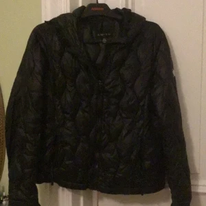 Svart quiltad pufferjacka från Amisu - Svart pufferjacka från Amisu i storlek 38. Jackan har ett quiltat mönster, huva och dragkedja framtill. Perfekt för kyliga dagar och har en snygg, lite boxig passform. Materialet är syntet och jackan är långärmad.
