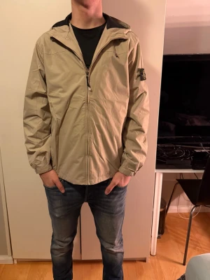 Beige vindjacka från Stone Island - Säljer en beige vindjacka från Stone Island med huva och dragkedja framtill. Jackan har den klassiska Stone Island-loggan på vänster ärm och är tillverkad i lätt polyester. Perfekt för dig som vill ha en clean och stilren look.