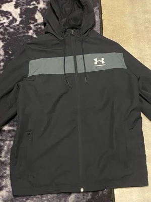 Svart vindjacka från Under Armour - Svart vindjacka från Under Armour med grått band över bröstet och logga. Jackan har huva med snörning, dragkedja framtill och två sidofickor. Perfekt för dig som gillar sportig stil och vill ha något lätt att slänga på sig.