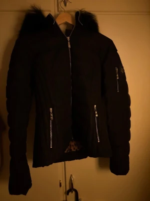 Svart pufferjacka från Hollies - Svart pufferjacka från Hollies med fluffig päls på huvan och leopardmönstrat foder. Jackan har flera silvriga dragkedjor, design och långärmade ärmar. Perfekt för dig som vill ha en snygg och varm jacka med coola detaljer.