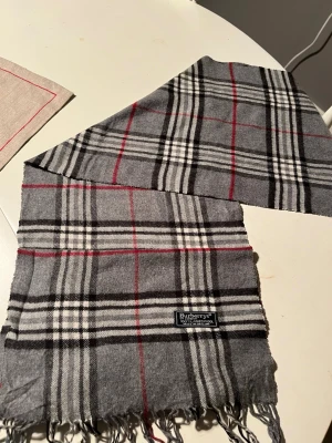 Grå burberry scarf - Säljer en helt oanvänd scarf från burberry, scarfen har ett rutigt mönster och fransar på sidorna. Den är gjord på 100% lammull och är väldigt skön. Priset är ej hugget i sten!