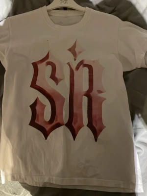 Vit t-shirt med röd textprint - Vit t-shirt från Biot med stor röd textprint i gotisk stil på både fram- och baksidan. Klassisk rund hals och korta ärmar. Tillverkad i mjuk bomull som känns skön mot huden. Perfekt för dig som gillar streetwear och grafiska tryck.