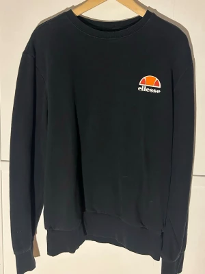 Svart sweatshirt från Ellesse XL - Svart sweatshirt från Ellesse i storlek XL. Tröjan har rund hals, långa ärmar och ett broderat Ellesse-logo på bröstet i orange, rött och vitt. Mjuk och skön bomullskänsla med ribbade muddar vid ärmslut och nederkant.