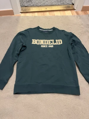 Mörkgrön sweatshirt från Bondelid S - Säljer en mörkgrön sweatshirt från Bondelid med beige text 'BONDELID SINCE 1956' på bröstet. Klassisk rund halsringning och långa ärmar. Tillverkad i mjuk bomull för skön vardagsstil.