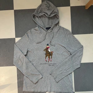 Grå hoodie Polo Bear Ralph Lauren - Grå hoodie från Polo Ralph Lauren med  Polo Bear-tryck på bröstet där björnen rider häst. Tröjan har huva med snörning och är tillverkad i mjuk bomull. Långärmad och med avslappnad passform, perfekt för en chill och stilren look. Är äkta såklart! Personen på bilden är 180 cm lång och väger 70 kg. Pris kan bli diskuterad! 
