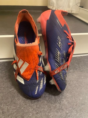 Adidas Predator blå/orange fotbollsskor - Säljer ett par Adidas Predator fotbollsskor i blått och orange med vita detaljer. Skorna har snörning, låg profil och mönstrad ovandel för extra grepp på planen. Sulan har fasta dobbar och insidan är vadderad för skön passform. Perfekta för dig som vill sticka ut på matchen!